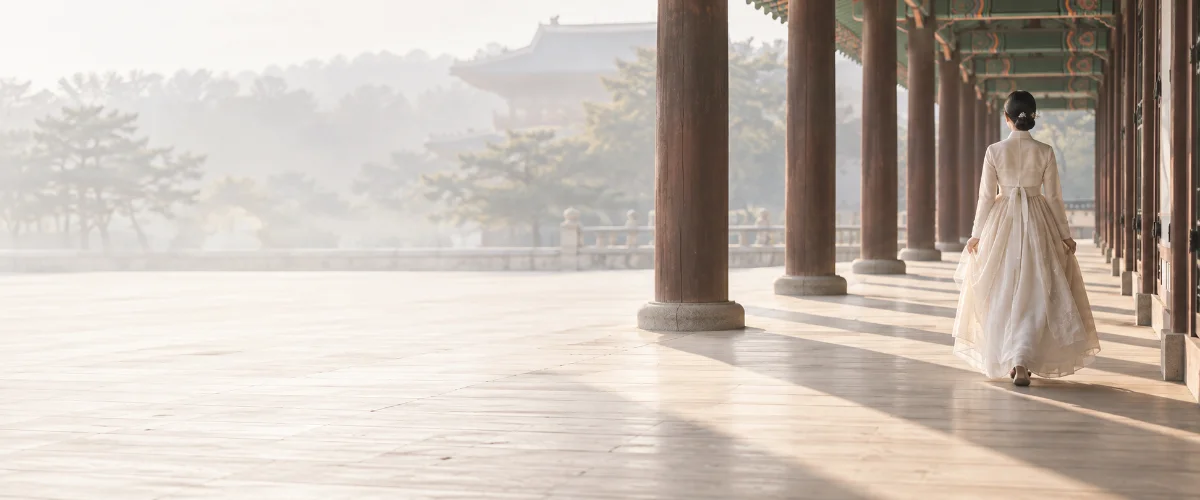 A woman in hanbok walking along a palace corridor in the morning light